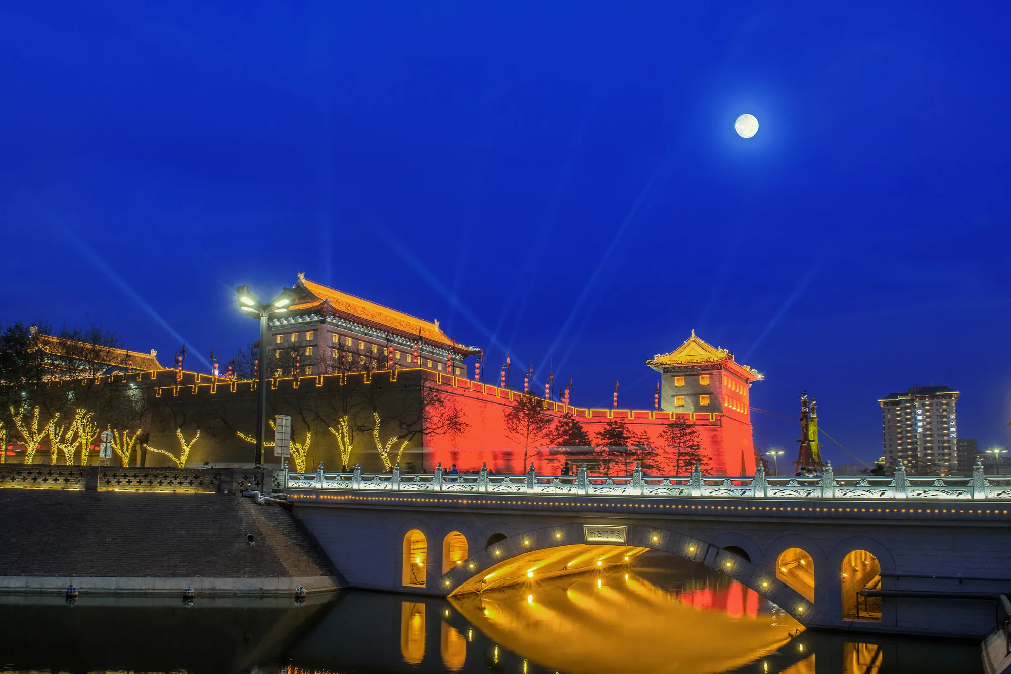 Xi'an City Wall illuminated red and gold above its moat beneath a full moon at night