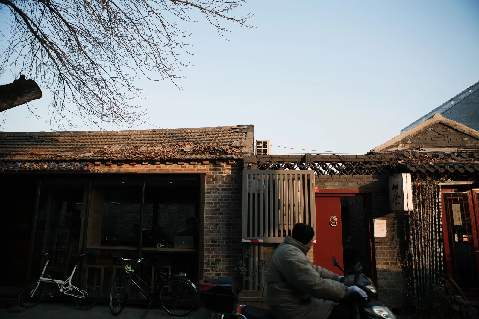 A scooter rider passing bicycles, old brick courtyards and a coffee shop in a lived-in Beijing hutong